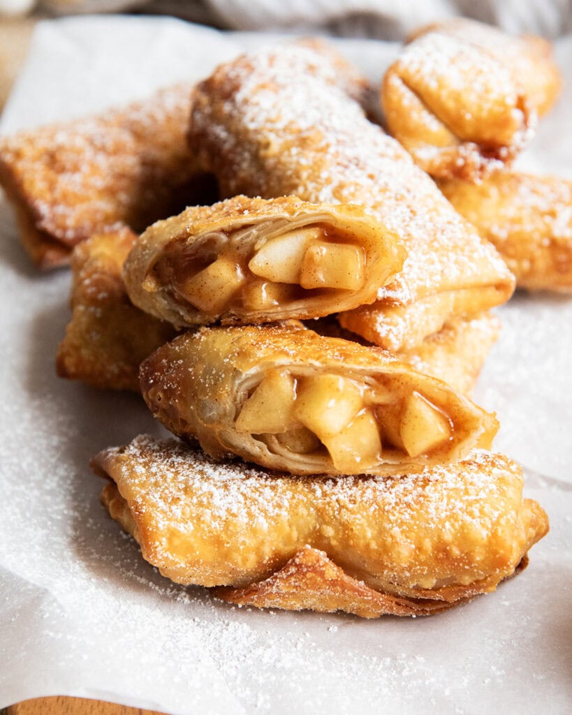 A pile of apple pie egg rolls, two are cut in half showing the apple pieces in the middle.