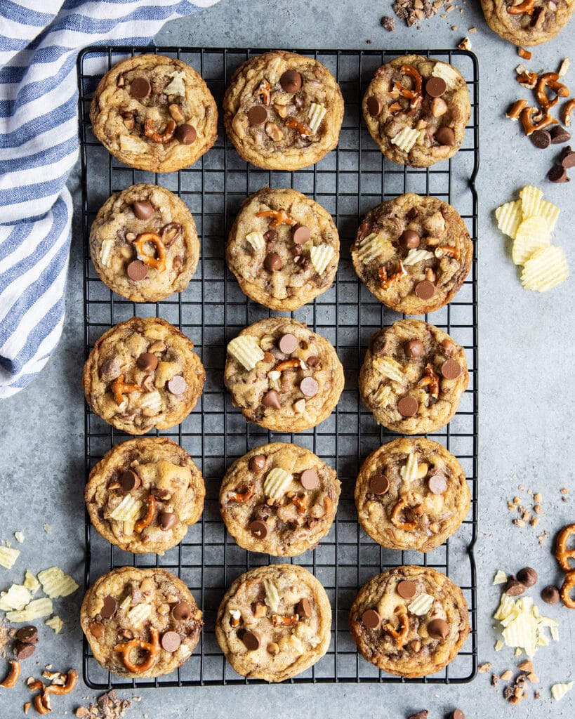 An above view of sweet and salty cookies on a cooling rack with pretzels, potato chips, and chocolate chips.