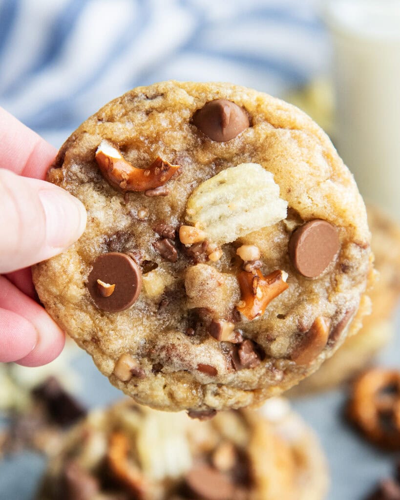 A hand holding a chocolate chip cookie with pretzel pieces, and chip pieces in it.