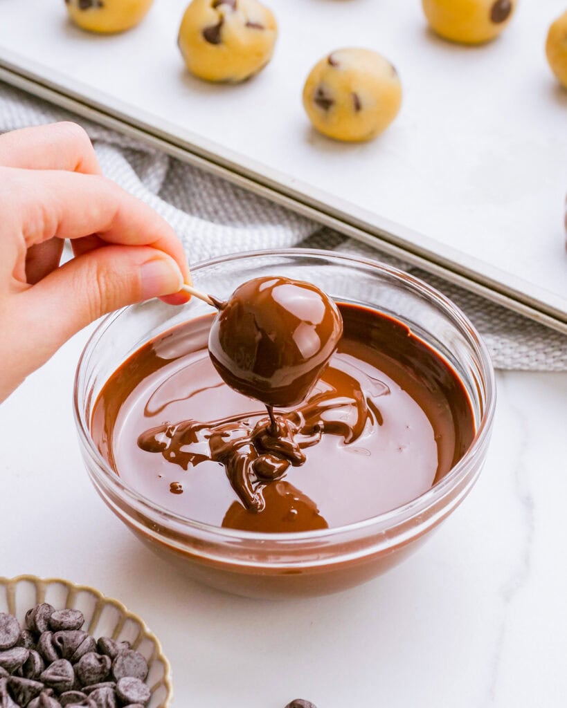 A cookie dough ball dipped in chocolate on a toothpick above a bowl of melted chocolate. 