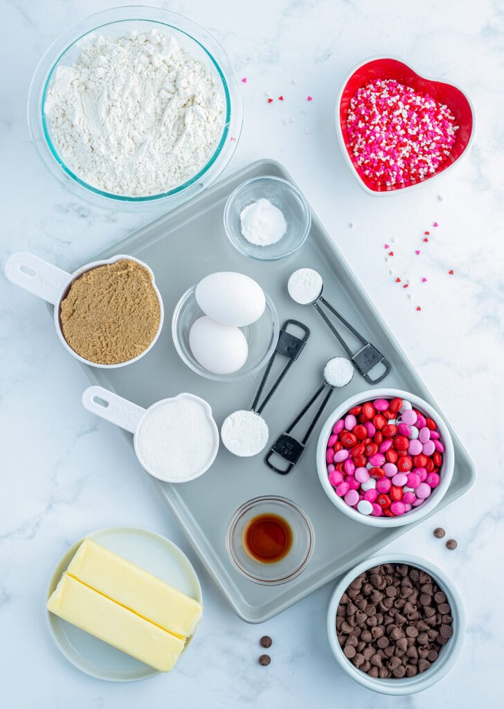 The ingredients needed to make Valentine M&M cookies on a baking sheet, butter, sugar, chocolate chips, m&ms, flour, sprinkles, and more. 