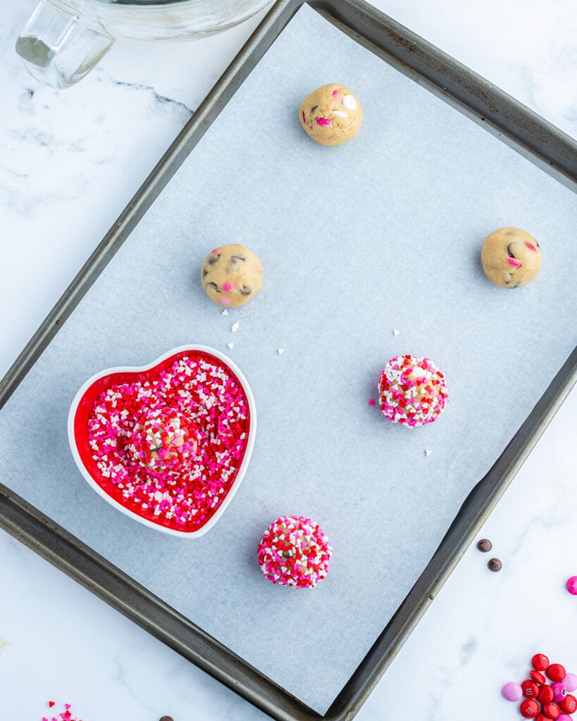 Cookie dough balls on a baking sheet, three of them are rolled in valentine sprinkles.