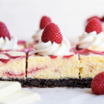 A side view of Cheesecake bars on a piece of parchment paper, topped with whipped cream, and fresh raspberries.