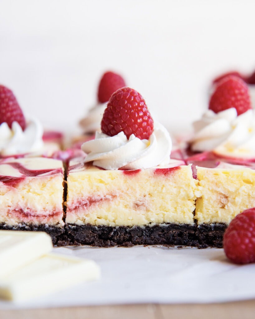 A side view of Cheesecake bars on a piece of parchment paper, topped with whipped cream, and fresh raspberries. 