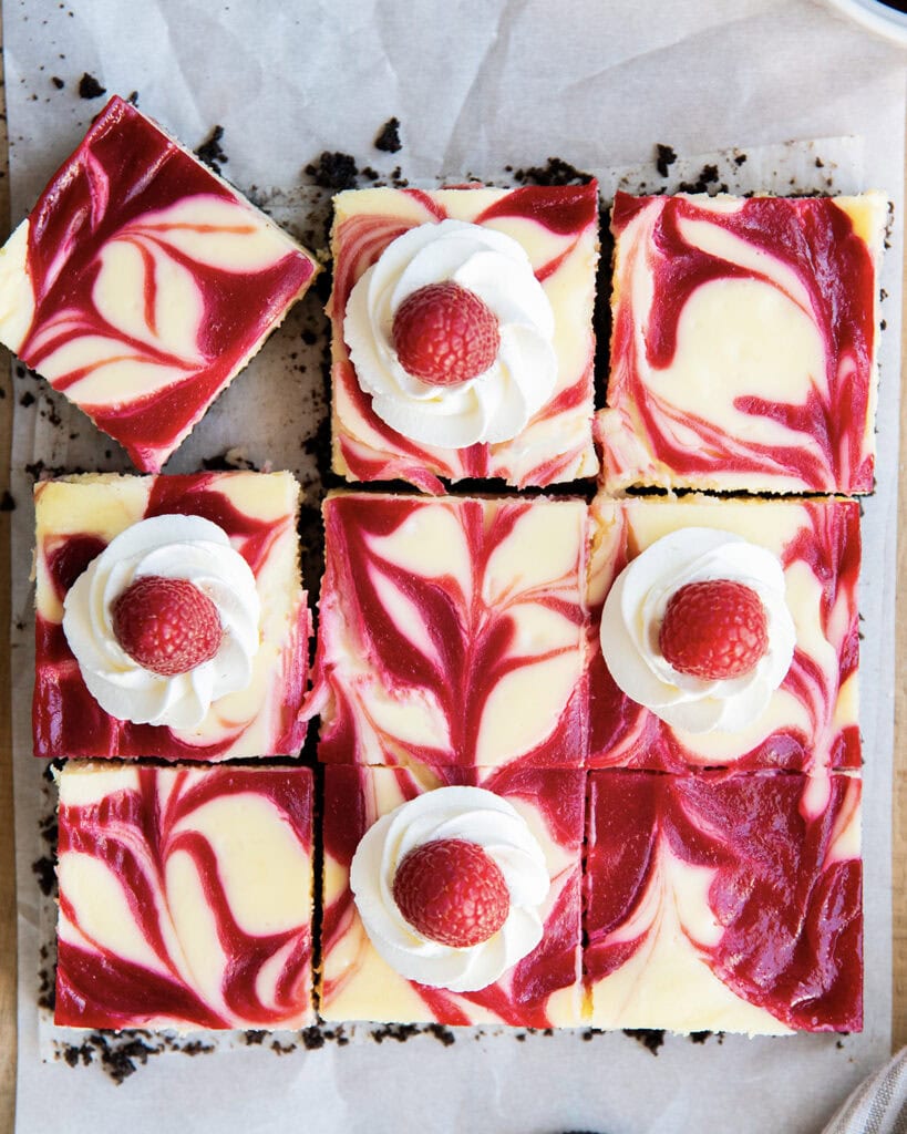 An above view of squares of raspberry cheesecake with raspberry swirls and whipped cream dollops on top.