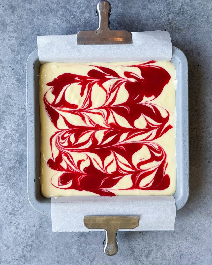 An above view of an uncooked raspberry swirled cheesecake in a square pan.