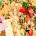 A chip dipping into a pan of beef queso topped with tomatoes, and cilantro.