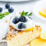 A piece of lemon ricotta cake on a plate topped with a dollop of whipped cream and fresh blueberries.
