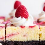 A side view of cheesecake bars with an Oreo crust, the cheesecake has a swirl of raspberry through it, and are topped with whipped cream and a fresh raspberry.