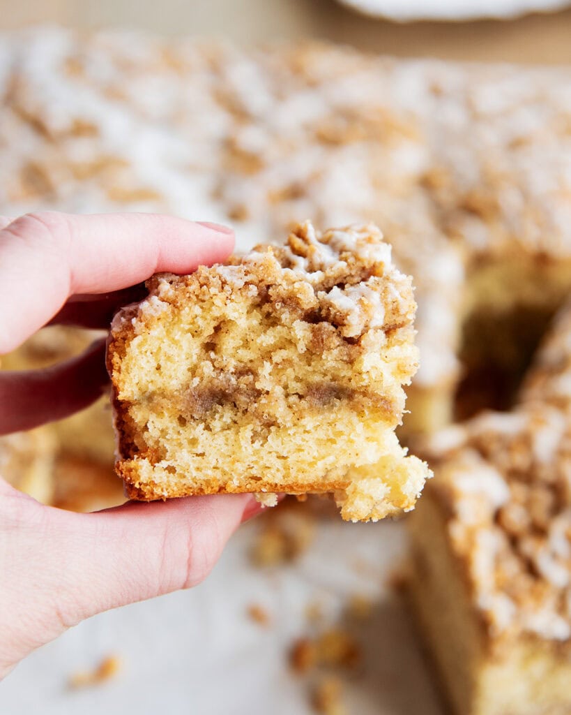 A hand holding a soft piece of cinnamon coffee cake with a crumb topping.