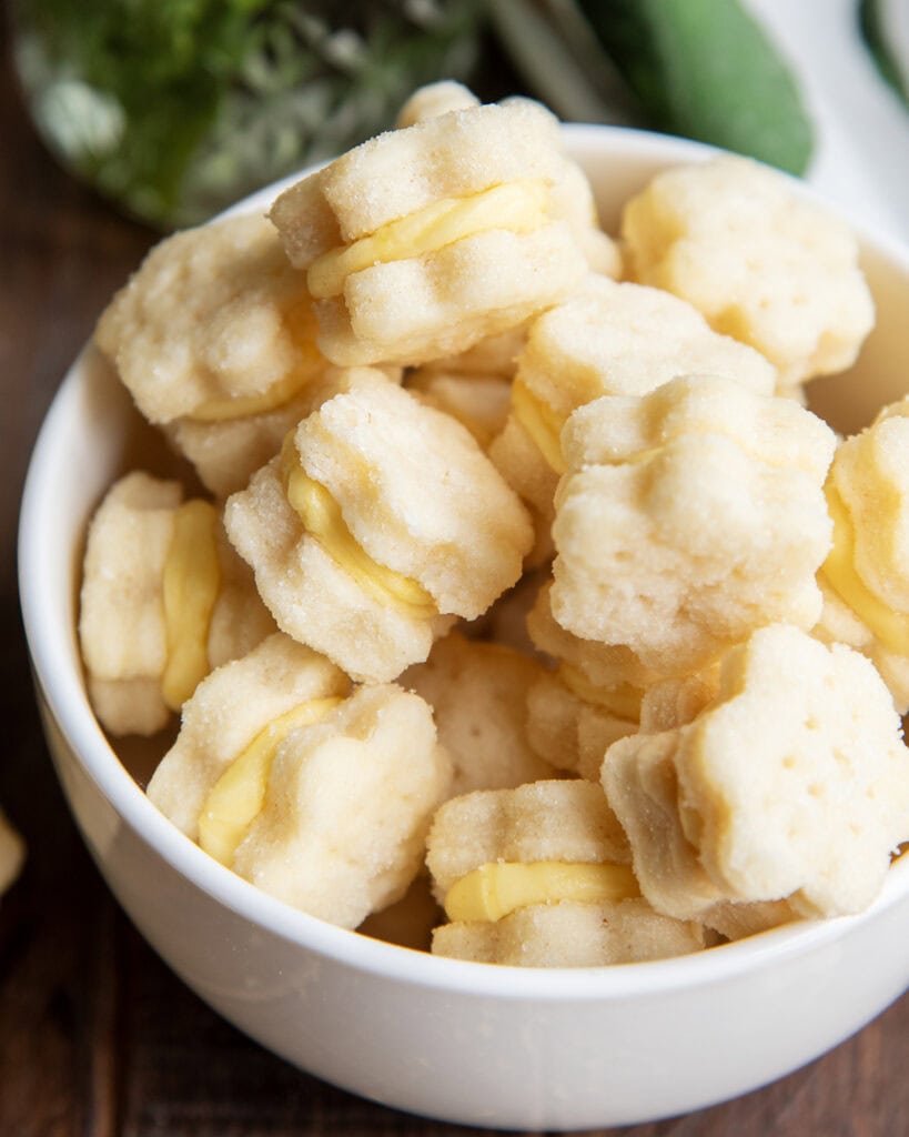 A close up of bowl of tiny flower shaped lemon cream wafer cookie sandwiches with a yellow lemon frosting.