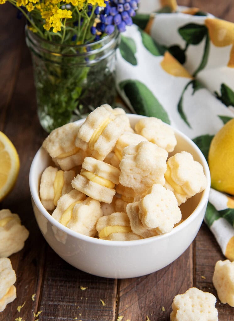 A bowl of tiny flower shaped cookie sandwiches with a yellow lemon frosting.