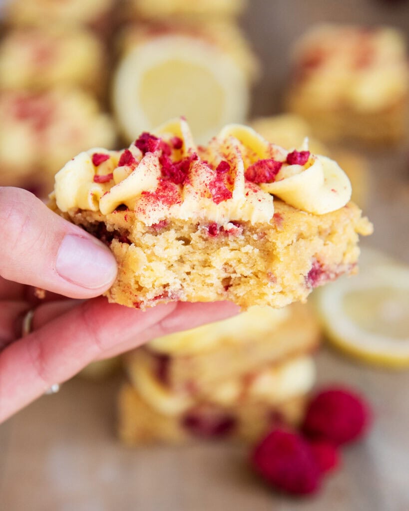 A hand holding a lemon raspberry blondie topped with lemon buttercream and freeze dried raspberries.