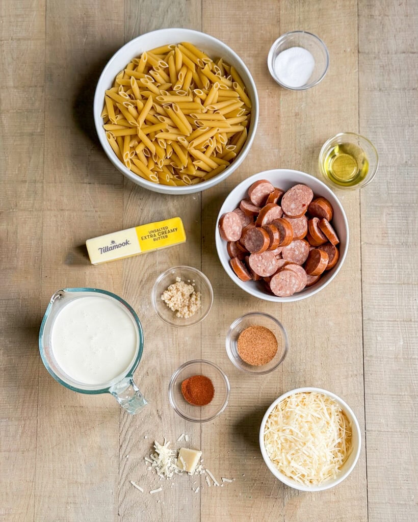 The ingredients needed to make sausage alfredo pasta including penne pasta, sausage slices, butter, cream, parmesan, cajun seasoning, and paprika. 