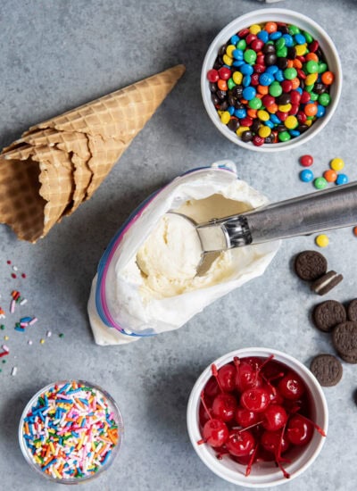 An above view of a scoop of ice cream on a scoop, in a ziplock bag, surrounded by ice cream toppings.