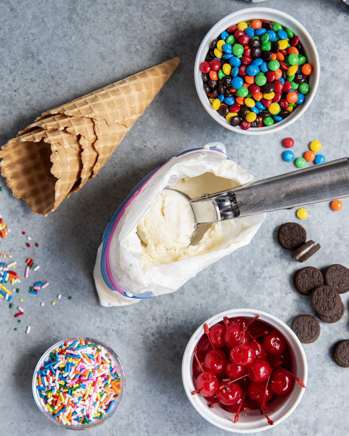 An above view of a scoop of ice cream on a scoop, in a ziplock bag, surrounded by ice cream toppings.