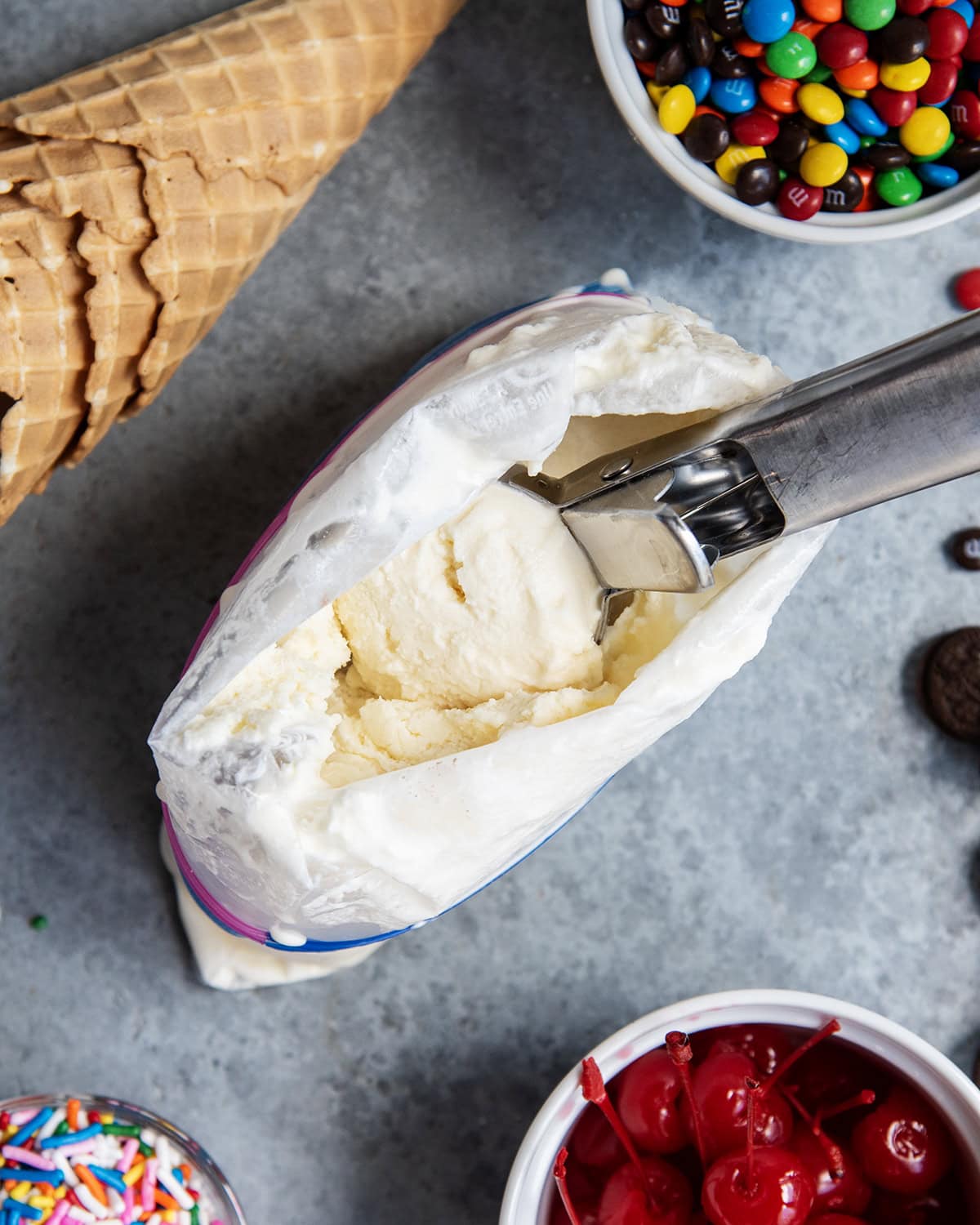 An above view of a scoop of ice cream on a scoop, in a ziplock bag, surrounded by ice cream toppings.
