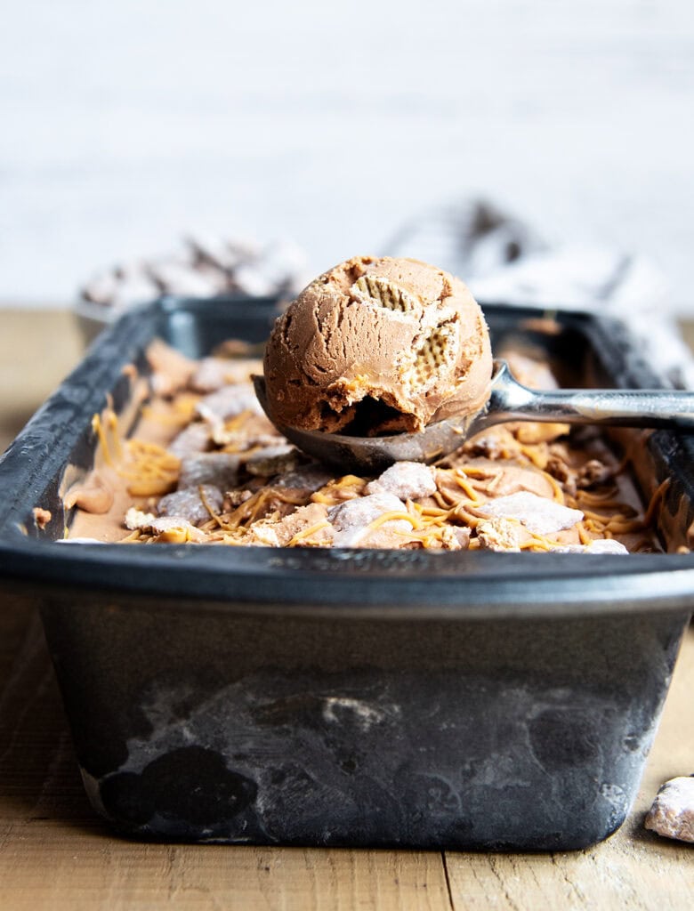 A scoop of chocolate ice cream with chex pieces and peanut butter swirled in it, on top of a container of Muddy Buddy Ice Cream.
