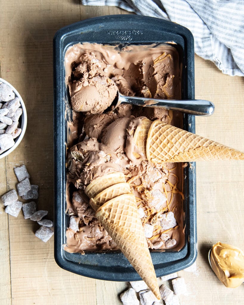 An above view of a container of chocolate muddy buddy ice cream, with two ice cream cones of it on top.