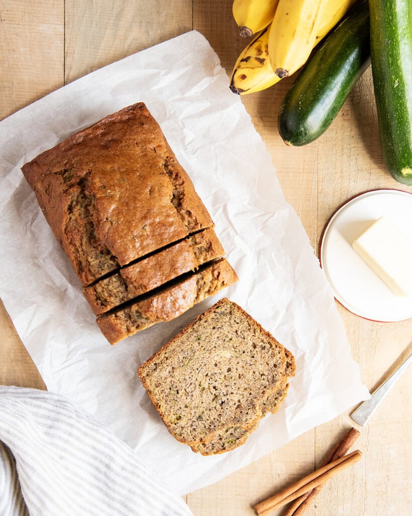 An above view of a loaf of banana zucchini bread, with slices cut off, and laying in front on a piece of white parchment paper.
