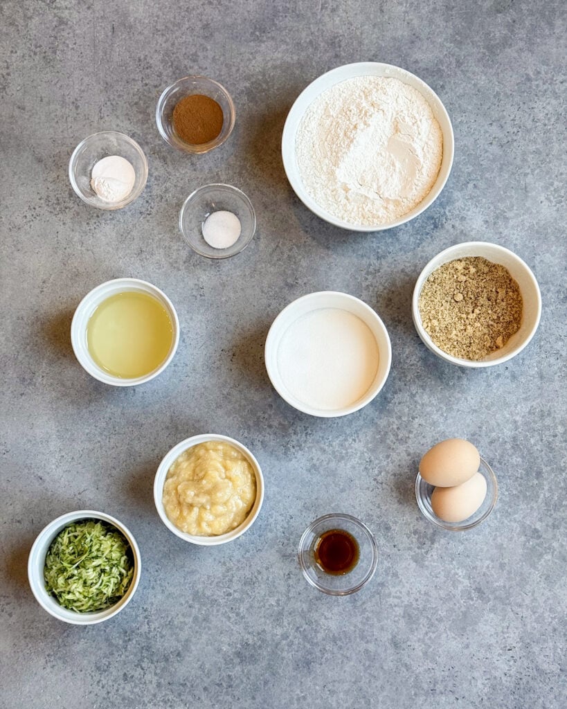 Small bowls of the ingredients needed to make banana zucchini bread, including flour, cinnamon, zuccini, banana, eggs, oil, sugar, and more.