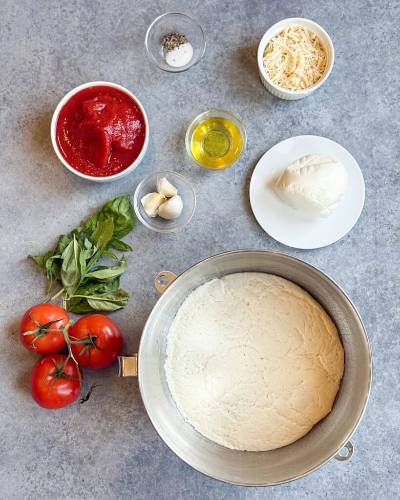 The ingredients needed to make margherita pizza including dough, sauce, fresh mozzarella, basil, and more.