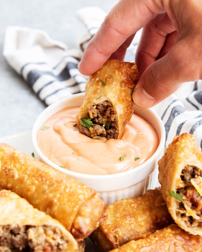 A hand holding half a cheeseburger egg roll and dipping it into fry sauce.
