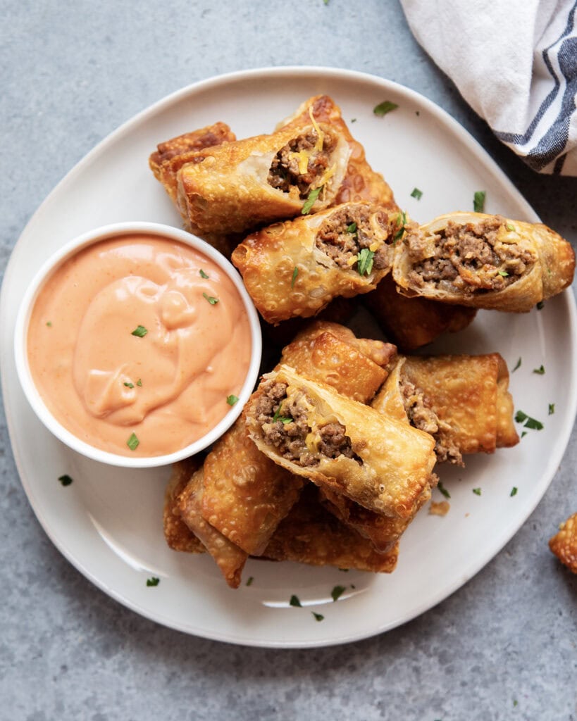 An above view of cheeseburger egg rolls on a plate with a bowl of fry sauce.