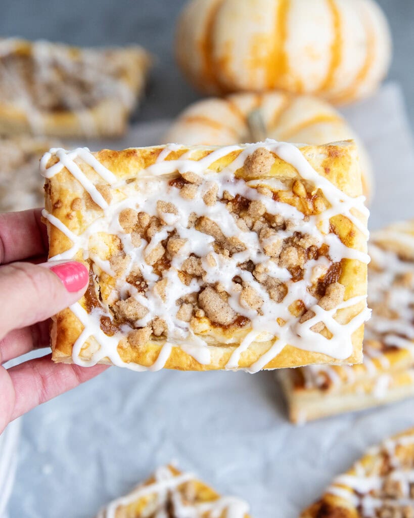 A hand holding a cream cheese danish topped with a crumble topping and vanilla icing.