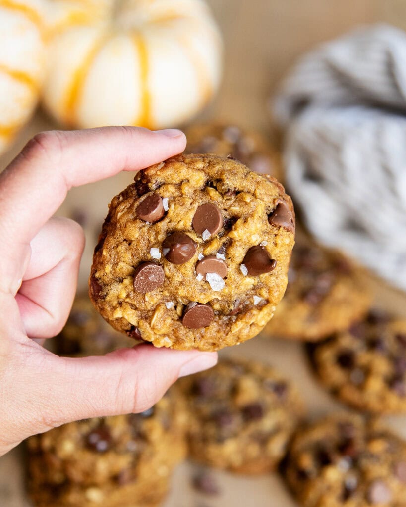 A hand holding a pumpkin chocolate chip cookie with melty chocolate chips and sea salt on top.