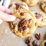 A hand holding a butter pecan cookie with a pecan half and flaky sea salt on top.