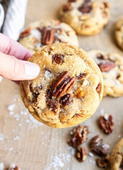 A hand holding a butter pecan cookie with a pecan half and flaky sea salt on top.