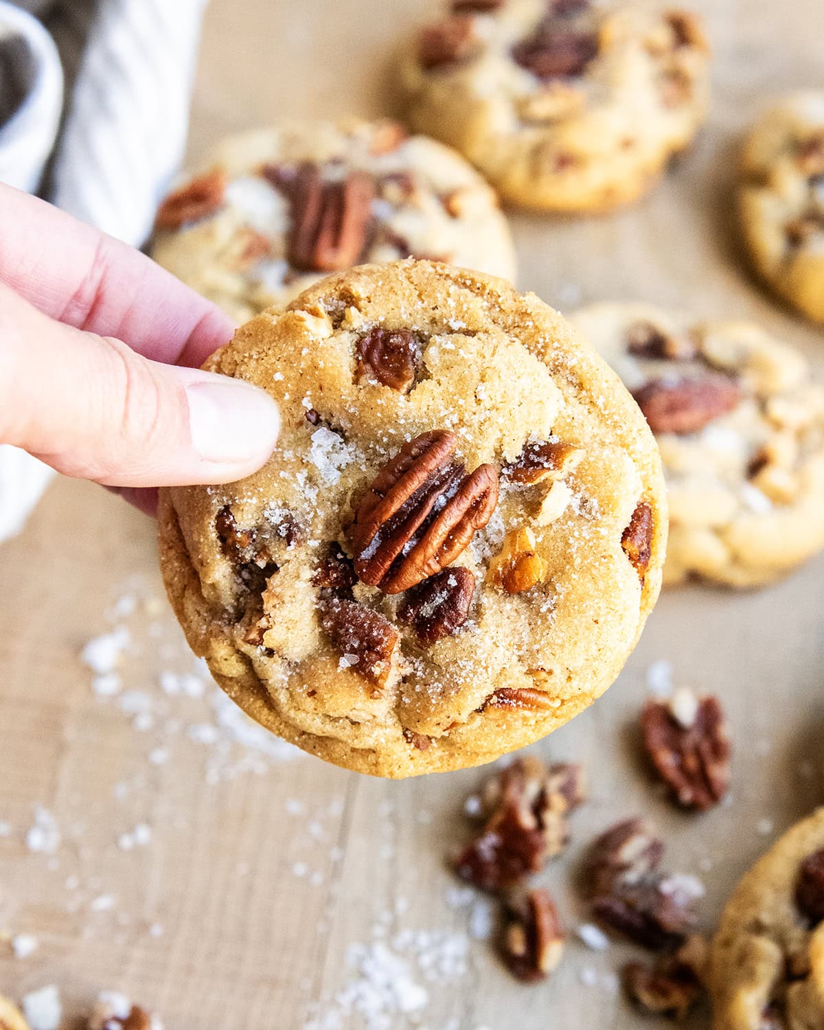 A hand holding a butter pecan cookie with a pecan half and flaky sea salt on top.