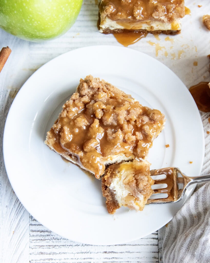 A caramel apple cheesecake bar on a plate with a forkful of it next to the bar. 