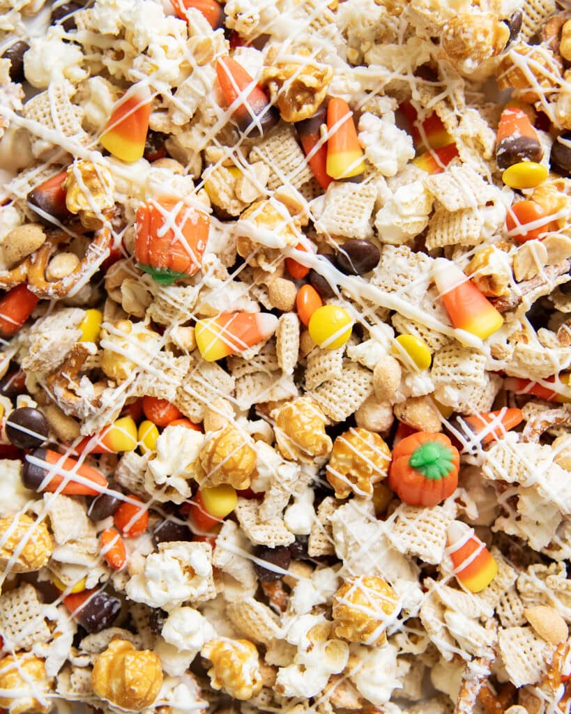 A close up of halloween snack mix, with white chocolate coated chex and popcorn, and reeses pieces and candy corn.