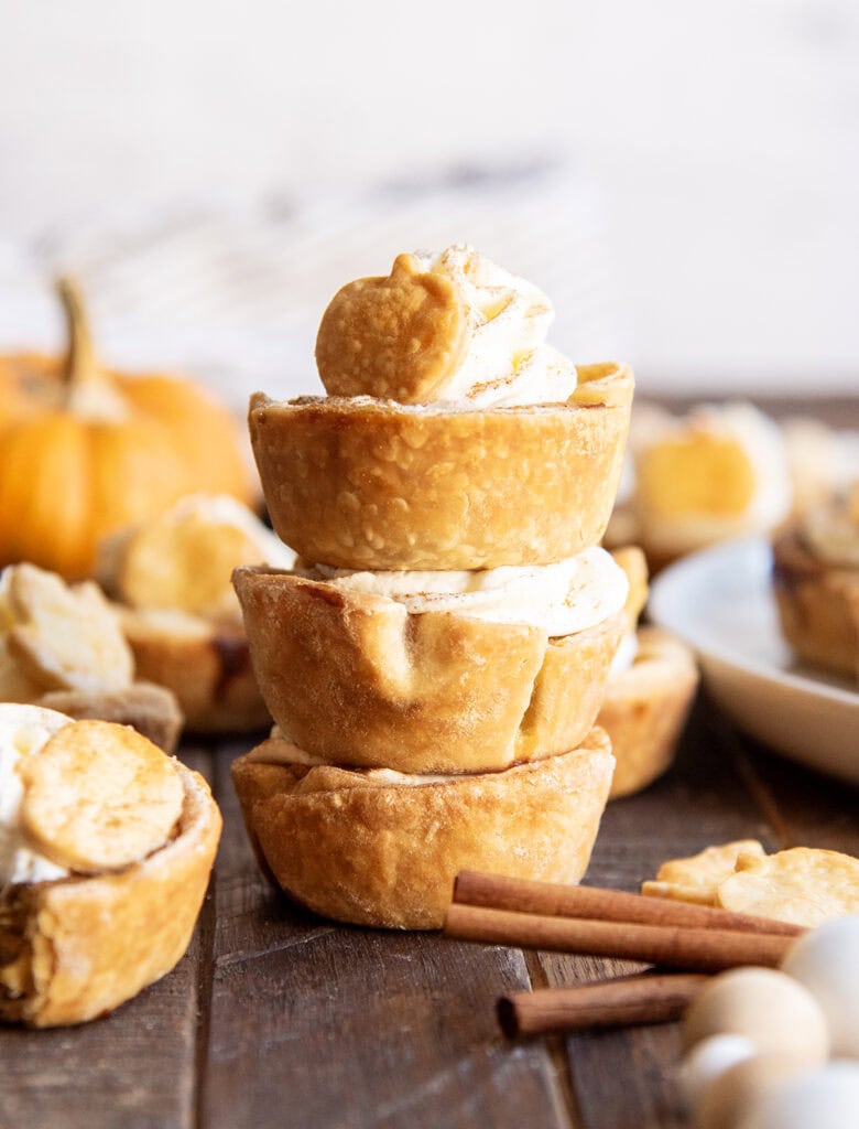 A stack of three mini pumpkin pies, the top one is topped with whipped cream and a pie crust topper.