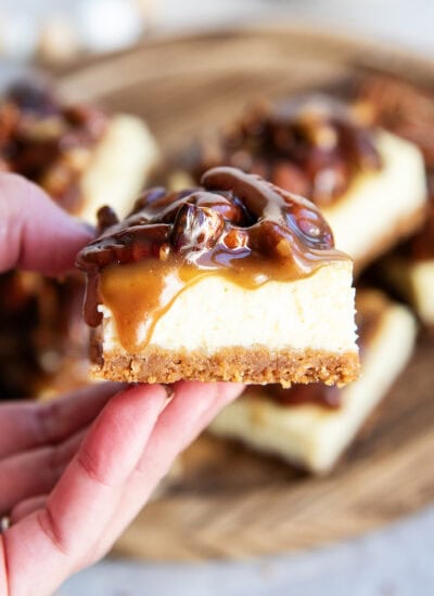A pecan pie cheesecake bar, topped with caramel and pecan topping, being held in a hand.