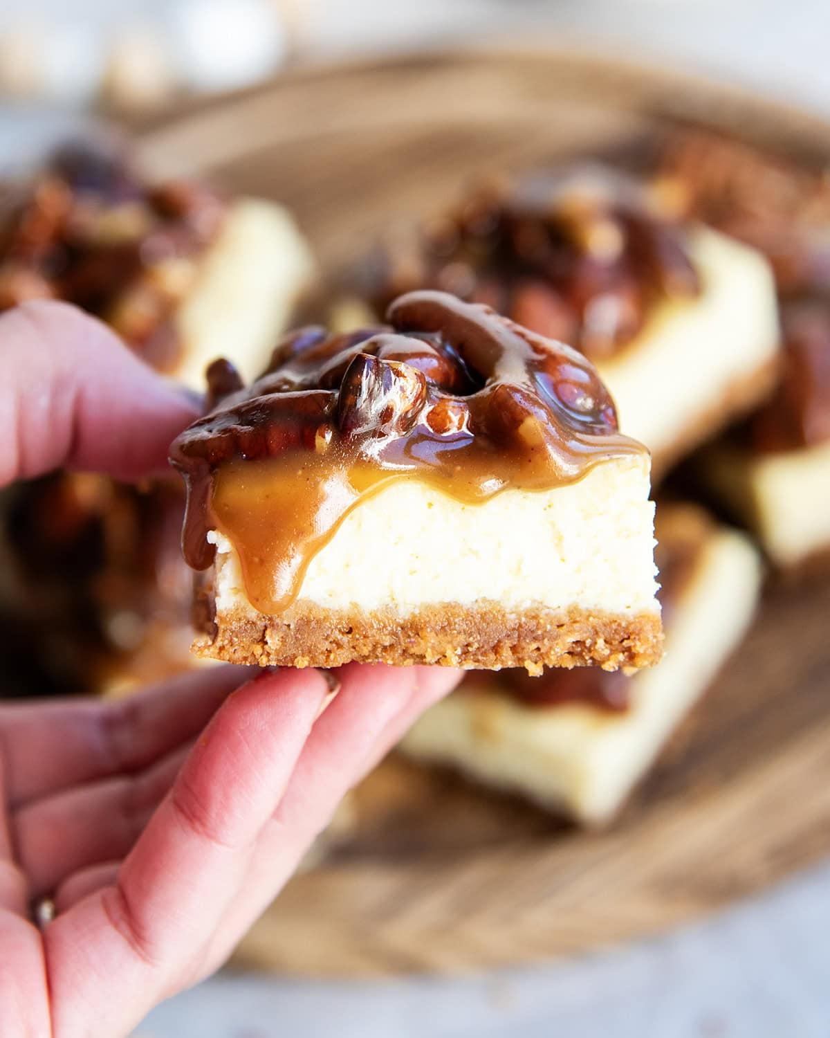 A pecan pie cheesecake bar, topped with caramel and pecan topping, being held in a hand.