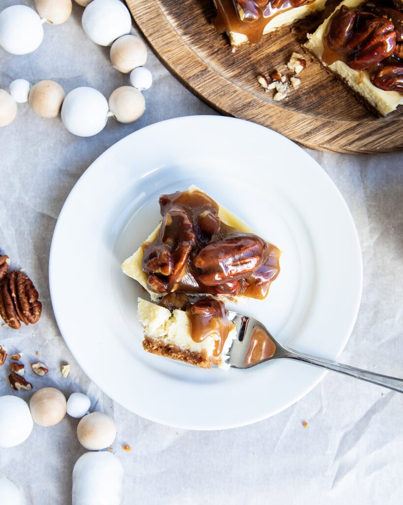 A plate with a pecan pie cheesecake bar with a piece cut out on a fork. 