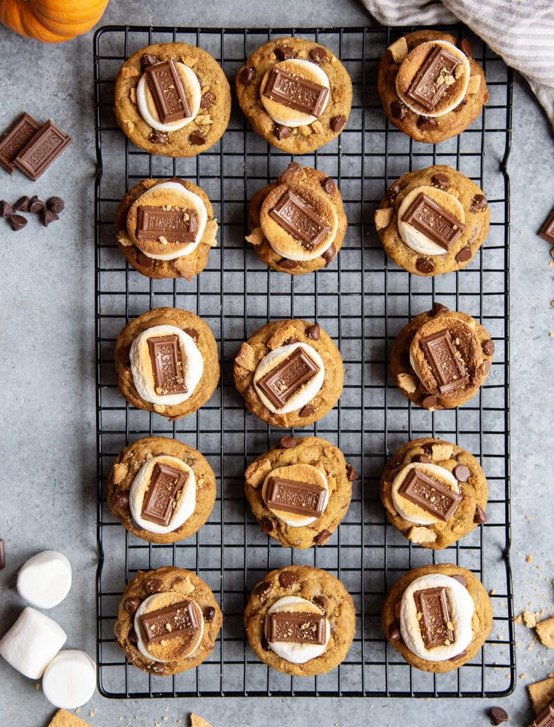 An above view of a cooling rack of pumpkin smores cookies with marshmallows and chocolate on top.