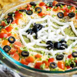 A glass dish filled with seven layer dip topped with a sour cream piped spider web and olive spiders.