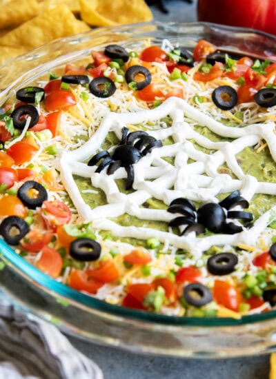 A glass dish filled with seven layer dip topped with a sour cream piped spider web and olive spiders.