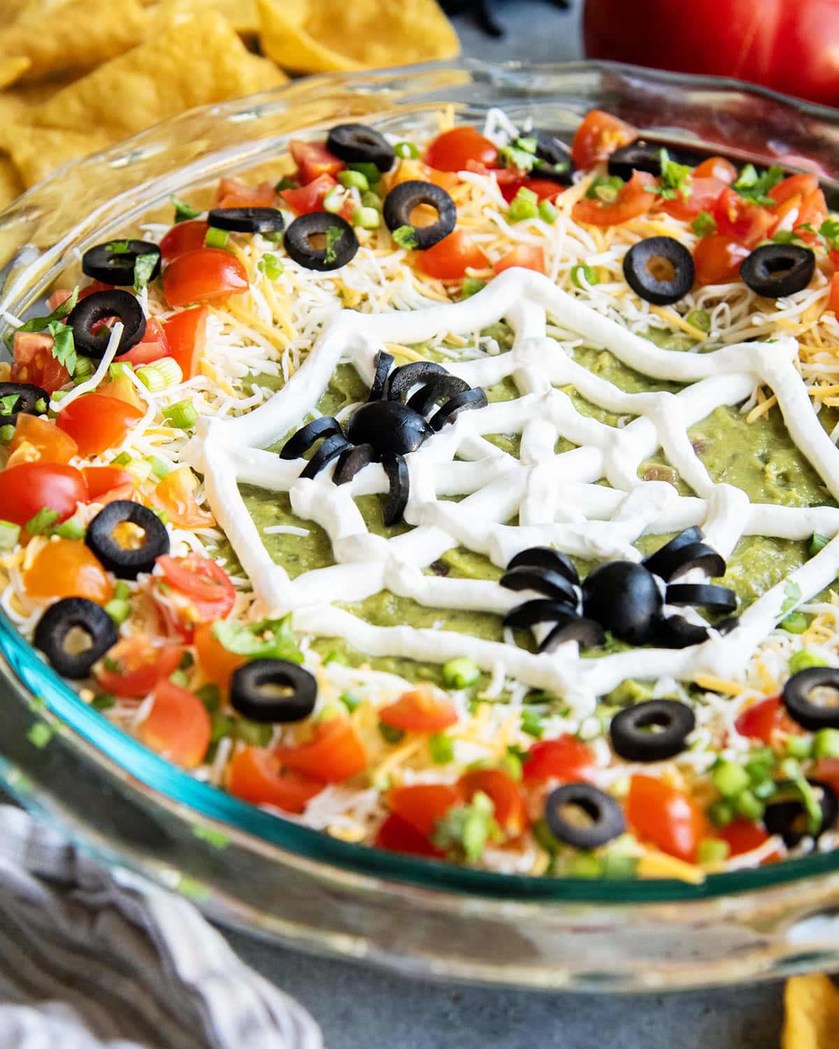 A glass dish filled with seven layer dip topped with a sour cream piped spider web and olive spiders.