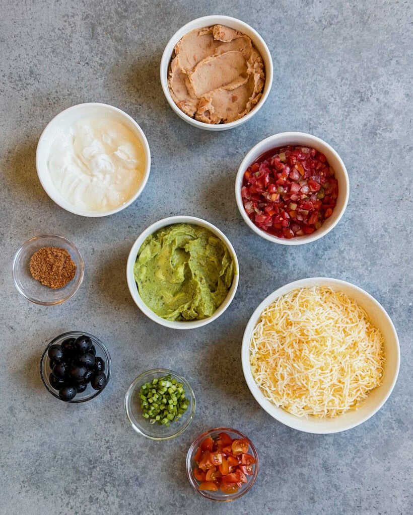 Small bowls of the ingredients needed to make seven layer dip, including sour cream, salsa, cheese, taco seasoning, and refried beans.