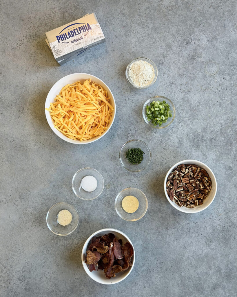 The ingredients needed to make a bacon ranch cheese ball in small bowls, like cream cheese, cheddar cheese, bacon, ranch seasoning, and pecans. 