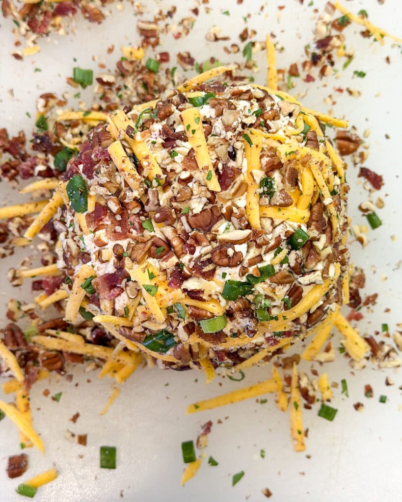 A cheese ball wrapped in cheese, and pecans on a cutting board.