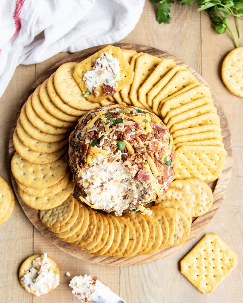 A cheeseball on a wooden platter from up above, it is surrounded by crackers, some have cream cheese on them. 