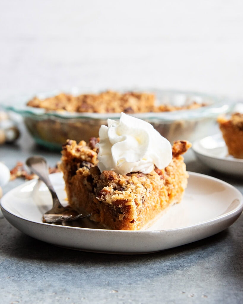 A slice of sweet potato pie with pecan topping on a plate with whipped cream on top.