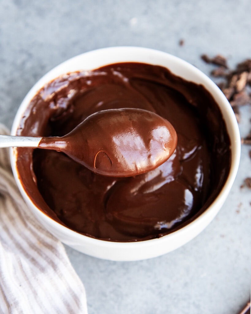 A bowl of chocolate ganache with a spoonful above it above the bowl.
