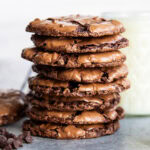 A stack of thin and chewy flourless chocolate cookies next to a cup of milk.